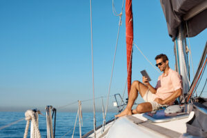 A white man in shorts and a T-shirt sits on the bow of his boat and uses a tablet