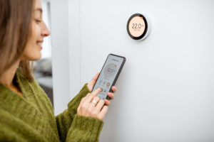 Woman stands in front of smart thermostat adjusting the temperature using her smartphone