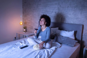 Woman sitting on bed eating popcorn and drinking wine while laughing at movie on TV